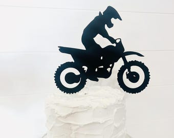 Peut inclure: Un topper de gâteau en forme de silhouette noire d'un motard de motocross sur une moto tout-terrain. Le topper est placé sur un gâteau glacé blanc.