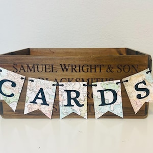 May include: A wooden card box with the text "SAMUEL WRIGHT & SON BLACKSMITHS" engraved on the front. A banner spelling "CARDS" hangs in front of the box. The banner flags have a vintage map design.