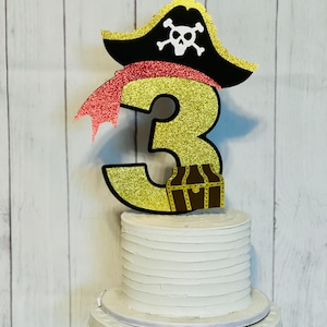May include: Gold glitter number three cake topper with a red glitter pirate hat and a brown treasure chest. The hat has a white skull and crossbones design.