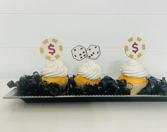 Peut inclure: Trois cupcakes jaunes avec un glaçage blanc surmontés de décorations sur le thème du casino. Un cupcake a une jeton de poker rouge et or avec un signe dollar, un autre a deux dés blancs avec des points noirs, et le troisième cupcake a une jeton de poker rouge et or avec un signe dollar. Les cupcakes sont sur un plateau argenté avec du papier déchiqueté noir.