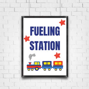 May include: A framed print with the words "FUELING STATION" in blue, with a colorful train illustration below. Red stars are scattered around the text. The train is red, yellow, and blue, with gray smoke coming from the engine.