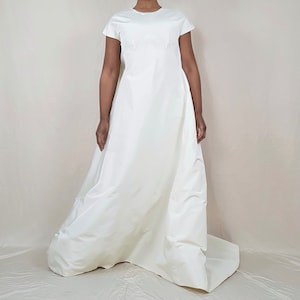 May include: A floor-length, ivory-colored wedding dress. The dress features a simple, round neckline and short sleeves. The skirt flows from the waist, creating a classic silhouette. The fabric appears to be a smooth, satin-like material.