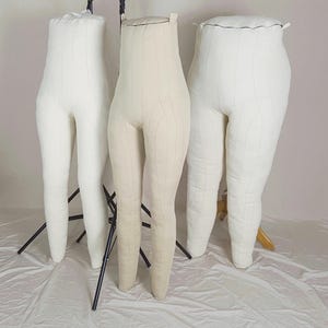 Puede incluir: Tres maniquíes acolchados en colores blanco roto y crema. Las formas tienen forma de piernas y están montadas sobre soportes de metal negro. Las formas están diseñadas para la confección y exhibición de prendas.