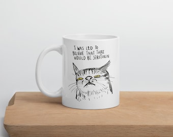 May include: White ceramic mug with a black and white illustration of a grumpy cat. The mug has the text "I WAS LED TO BELIEVE THAT THERE WOULD BE SEROTONIN" printed on it. The mug is sitting on a wooden surface.