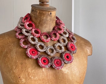May include: A multi-layered necklace made of crocheted fabric circles in shades of red, white, and brown. The circles are arranged in a cascading pattern, creating a bold and eye-catching statement piece.