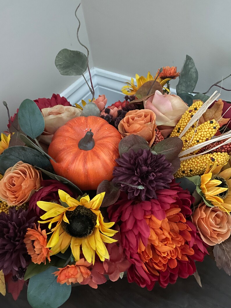 May include: A fall-themed centerpiece featuring a variety of flowers in shades of orange, red, and purple, including sunflowers, mums, and roses. A small orange pumpkin is nestled in the center of the arrangement.