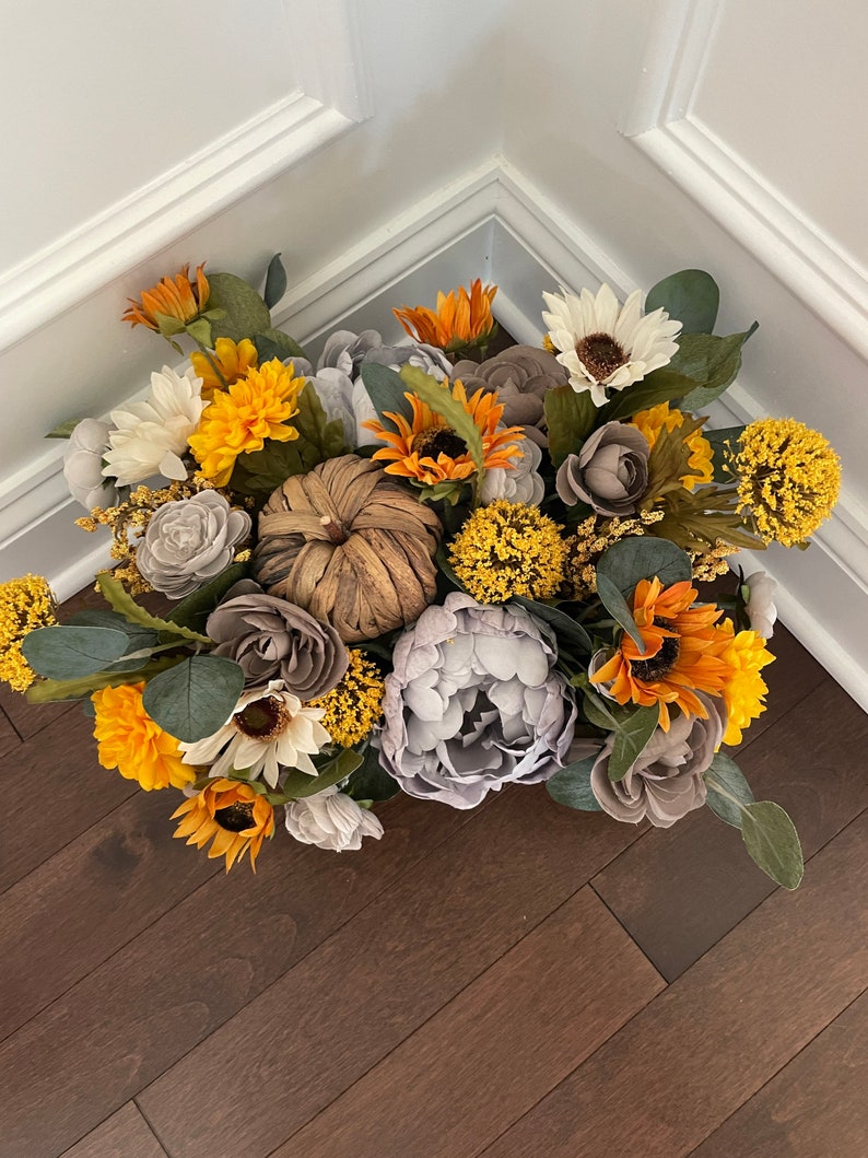 Peut inclure: Un arrangement floral sur le th&egrave;me de l'automne avec des tournesols, des pivoines grises et une petite citrouille rustique. L'arrangement est dans un r&eacute;cipient brun et est entour&eacute; de verdure.