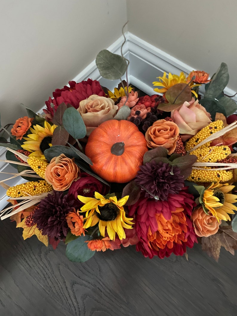 May include: A fall-themed floral arrangement featuring a variety of flowers in shades of red, orange, yellow, and brown. The arrangement includes sunflowers, roses, mums, and a small pumpkin.