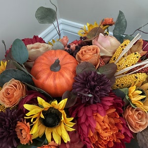 May include: A fall-themed centerpiece featuring a variety of flowers in shades of orange, red, and purple, including sunflowers, mums, and roses. A small orange pumpkin is nestled in the center of the arrangement.