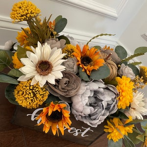 Peut inclure: Une bo&icirc;te en bois avec le mot "Harvest" &eacute;crit dessus, remplie d'un arrangement floral de tournesols, de fleurs blanches et grises, et de feuillage jaune et vert.