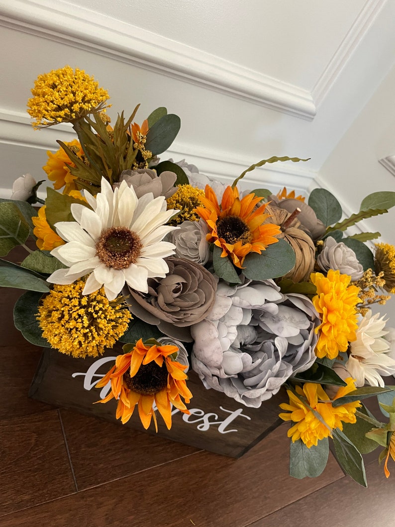 Peut inclure: Une bo&icirc;te en bois avec le mot "Harvest" &eacute;crit dessus, remplie d'un arrangement floral de tournesols, de fleurs blanches et grises, et de feuillage jaune et vert.