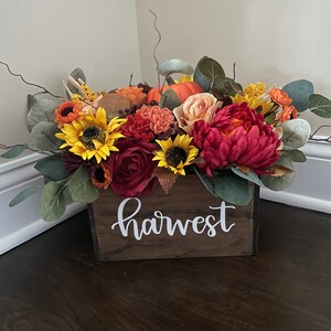 May include: A wooden box with the word "harvest" painted on the front, filled with a fall floral arrangement. The arrangement includes sunflowers, roses, and pumpkins in shades of orange, yellow, and red.