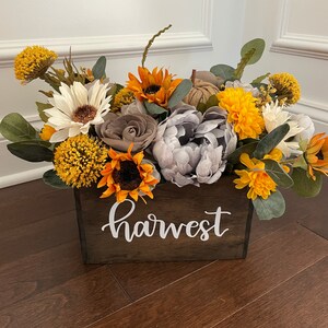 Peut inclure: Une bo&icirc;te en bois avec le mot "harvest" &eacute;crit dessus, remplie d'un arrangement floral de tournesols, de marguerites blanches, de pivoines grises et de chrysanth&egrave;mes jaunes.