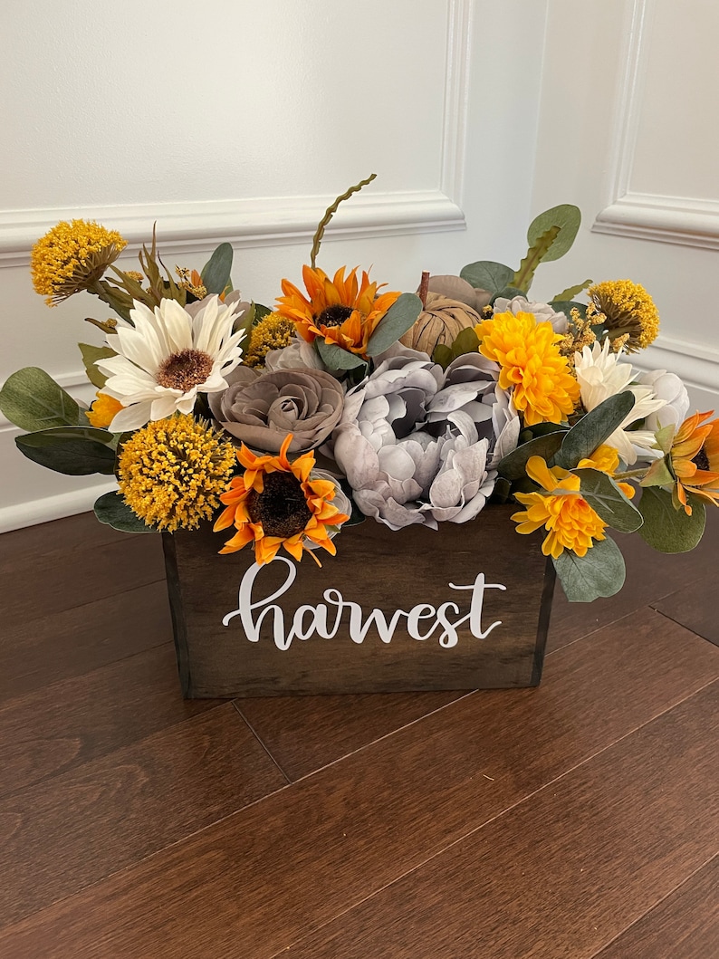 Peut inclure: Une bo&icirc;te en bois avec le mot "harvest" &eacute;crit dessus, remplie d'un arrangement floral de tournesols, de marguerites blanches, de pivoines grises et de chrysanth&egrave;mes jaunes.