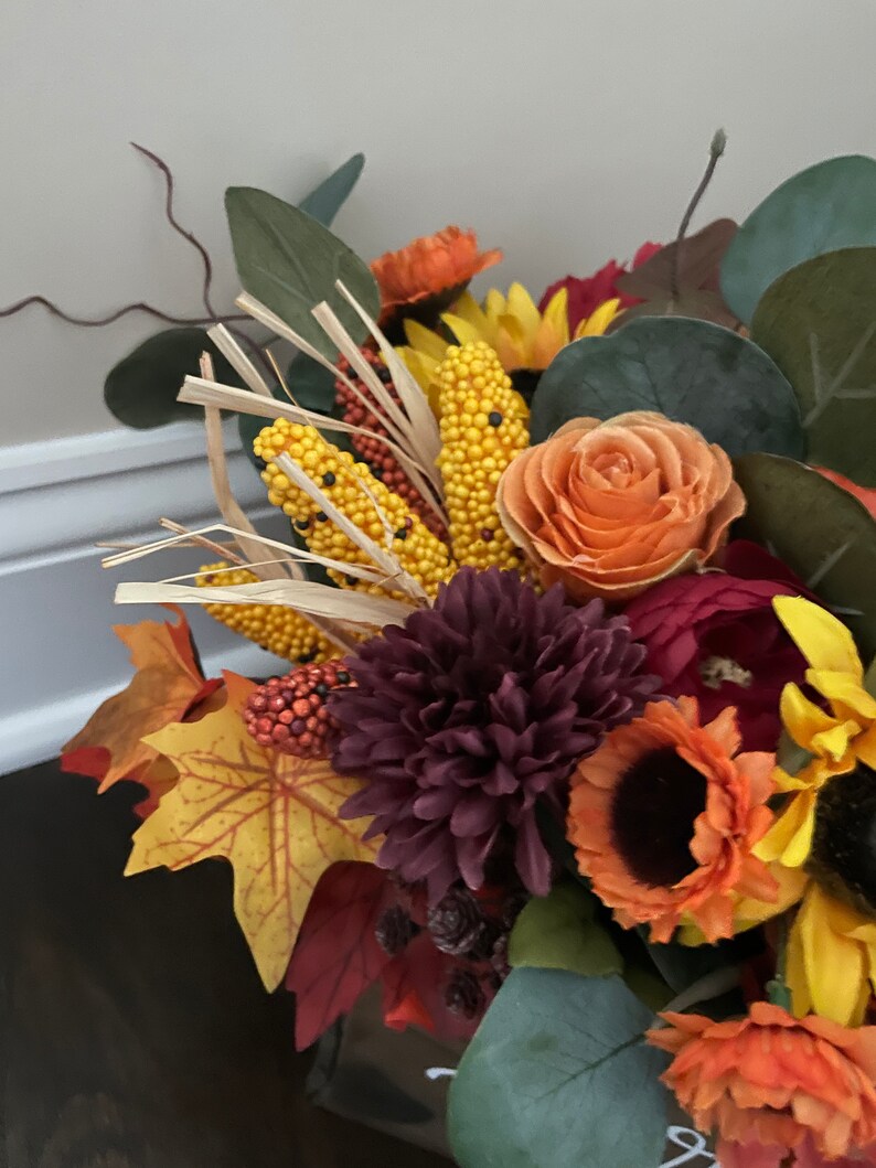 May include: A fall-themed floral arrangement with burgundy chrysanthemums, orange roses, sunflowers, and corn stalks. The arrangement is in a clear glass vase.