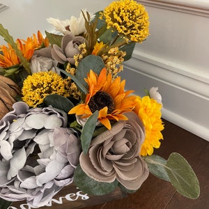 Peut inclure: Une bo&icirc;te en bois avec le mot "noves" &eacute;crit sur le devant, remplie d'un arrangement floral de tournesols, de pivoines grises et de chrysanth&egrave;mes jaunes.