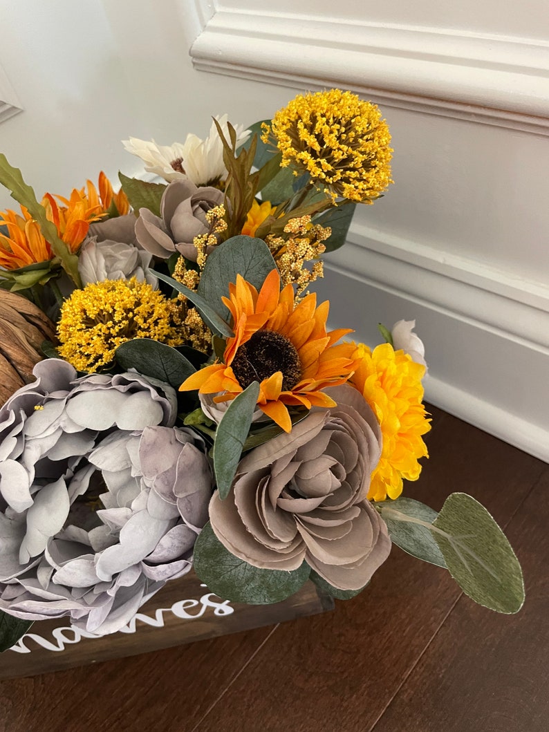 Peut inclure: Une bo&icirc;te en bois avec le mot "noves" &eacute;crit sur le devant, remplie d'un arrangement floral de tournesols, de pivoines grises et de chrysanth&egrave;mes jaunes.