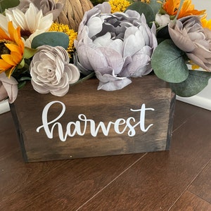 Peut inclure: Une bo&icirc;te en bois avec le mot "harvest" peint sur le devant. La bo&icirc;te est remplie d'un arrangement floral de tournesols, de pivoines et d'autres fleurs dans des tons de jaune, de gris et de brun.