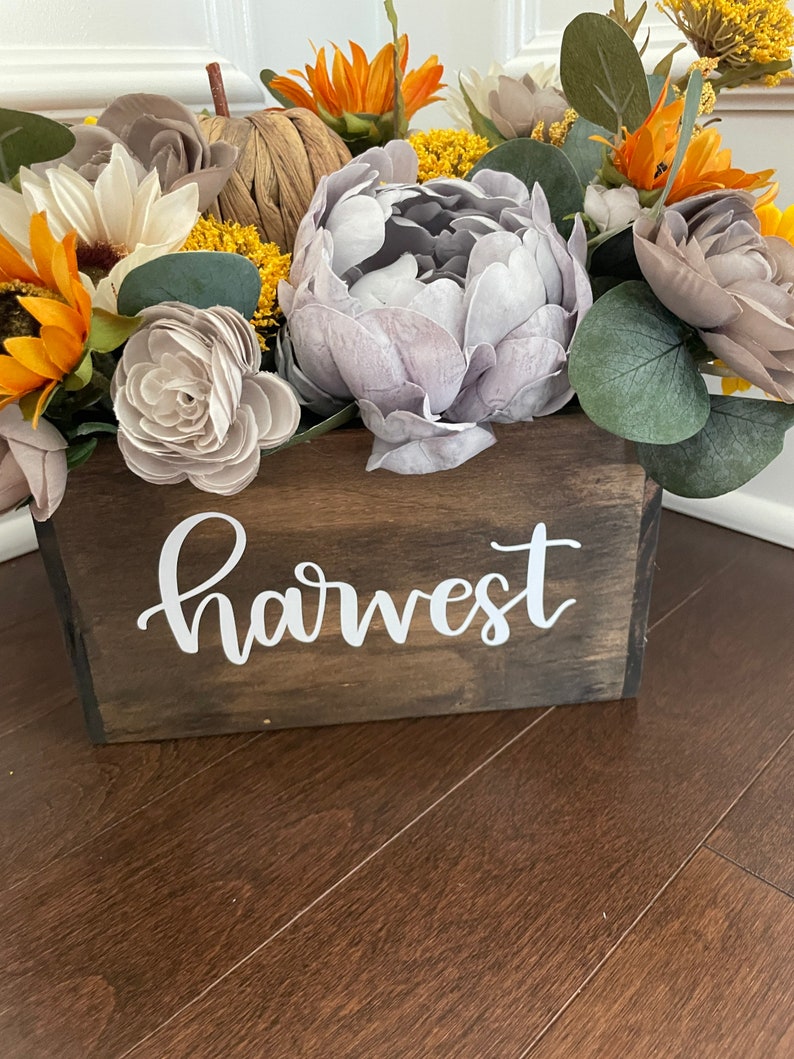 Peut inclure: Une bo&icirc;te en bois avec le mot "harvest" peint sur le devant. La bo&icirc;te est remplie d'un arrangement floral de tournesols, de pivoines et d'autres fleurs dans des tons de jaune, de gris et de brun.