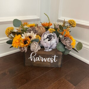 Peut inclure: Une bo&icirc;te en bois avec le mot "harvest" peint sur le devant, remplie d'un arrangement floral d'automne. L'arrangement comprend des tournesols, de l'eucalyptus et d'autres feuillages d'automne dans des tons de jaune, brun et gris.