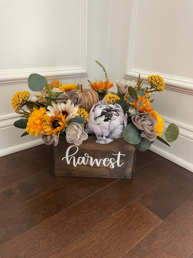 Peut inclure: Une bo&icirc;te en bois avec le mot "harvest" peint sur le devant, remplie d'un arrangement floral d'automne. L'arrangement comprend des tournesols, de l'eucalyptus et d'autres feuillages d'automne dans des tons de jaune, brun et gris.