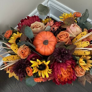 May include: A fall-themed floral arrangement featuring a variety of flowers in shades of red, orange, yellow, and brown. The arrangement includes sunflowers, roses, mums, and a small pumpkin.
