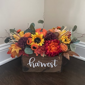 May include: A wooden box with the word "harvest" written on it. The box is filled with a fall floral arrangement featuring sunflowers, orange and red flowers, and a small pumpkin.