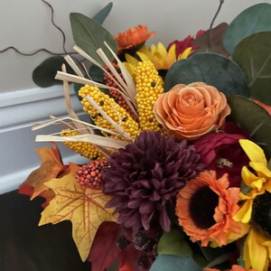 May include: A fall-themed floral arrangement with burgundy chrysanthemums, orange roses, sunflowers, and corn stalks. The arrangement is in a clear glass vase.