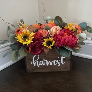 May include: A wooden box with the word "harvest" painted on the front, filled with a fall floral arrangement. The arrangement includes sunflowers, roses, and pumpkins in shades of orange, yellow, and red.