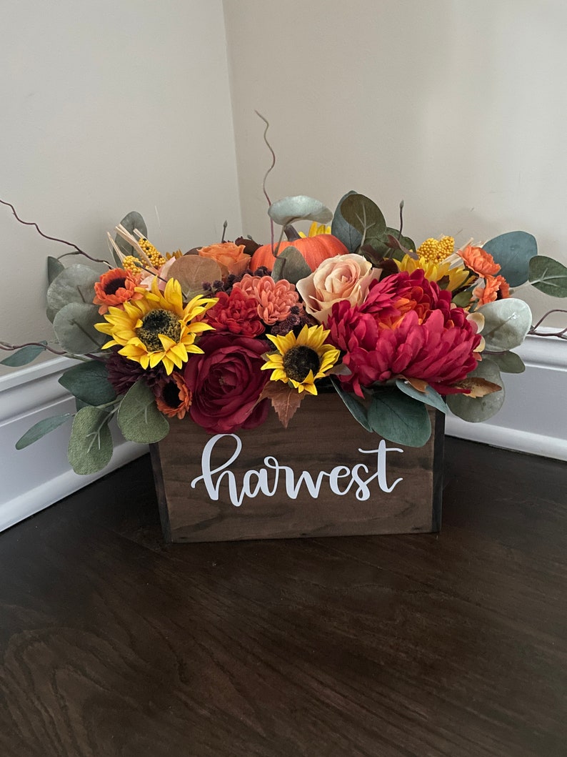 May include: A wooden box with the word "harvest" painted on the front, filled with a fall floral arrangement. The arrangement includes sunflowers, roses, and pumpkins in shades of orange, yellow, and red.
