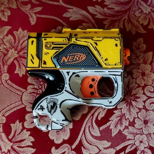 May include: A yellow, black, and white Nerf toy gun with orange detailing. The toy gun features the word "NERF". It is set against a red floral patterned backdrop.