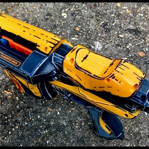 May include: A yellow and black toy blaster with orange accents. The toy gun has a futuristic design with the text "6 DARTS MAX" on the side. The blaster is lying on a textured gray surface.