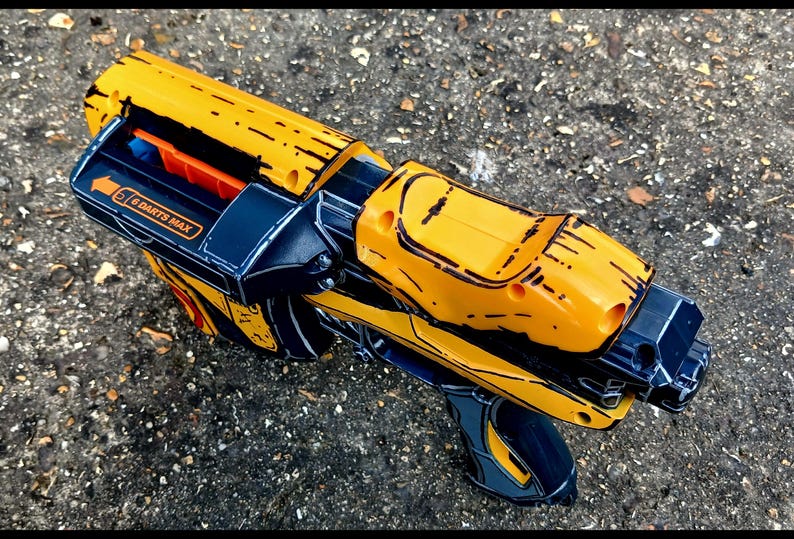 May include: A yellow and black toy blaster with orange accents. The toy gun has a futuristic design with the text "6 DARTS MAX" on the side. The blaster is lying on a textured gray surface.