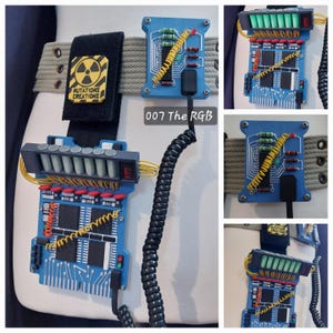 May include: A blue circuit board prop with a gray display and various colored wires and components. The prop is attached to a tan belt with a black strap featuring a yellow hazard symbol and the text "MUTATIONS CREATIONS". The text "007 The RGB" is also visible.