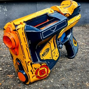 May include: A yellow, orange, and navy blue Nerf gun with a stylized design. The toy gun features the Nerf logo and a circular orange detail on the side. The gun has a trigger and a handle.