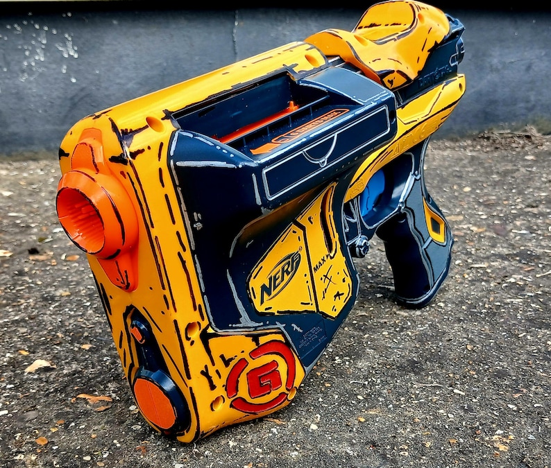 May include: A yellow, orange, and navy blue Nerf gun with a stylized design. The toy gun features the Nerf logo and a circular orange detail on the side. The gun has a trigger and a handle.