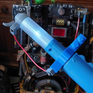 May include: A blue and white plastic toy proton pack with a red wire attached. The toy is partially assembled and is sitting on a table with a green ghost figure in the background.