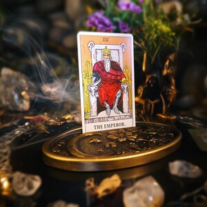 Op de afbeelding: Een tarotkaartlezing met een gele en witte kaart waarop de Keizer is afgebeeld, een man in een rode mantel die op een troon zit. De kaart ligt op een gouden metalen standaard met een maanontwerp. Er zijn kaarsen en rook te zien op de achtergrond.