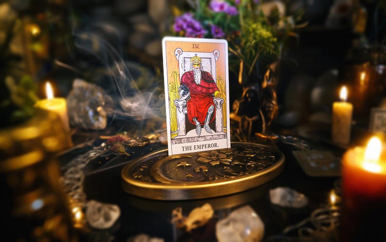 Op de afbeelding: Een tarotkaartlezing met een gele en witte kaart waarop de Keizer is afgebeeld, een man in een rode mantel die op een troon zit. De kaart ligt op een gouden metalen standaard met een maanontwerp. Er zijn kaarsen en rook te zien op de achtergrond.