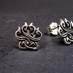 May include: A pair of silver-colored stud earrings. Each earring features a stylized, flame-like design with intricate, swirling details. The earrings are displayed on a dark, textured surface, highlighting their metallic sheen.