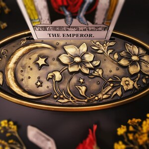 Op de afbeelding: Een gouden metalen dienblad met een sierlijk ontwerp met een halve maan, sterren en bloemmotieven. Het dienblad is ontworpen om een tarotkaart te houden, die zichtbaar is op de afbeelding en "THE EMPEROR." leest.