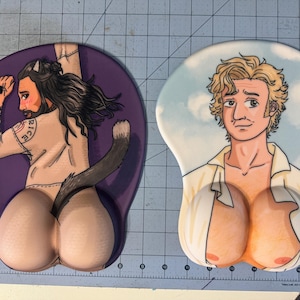 May include: Two mousepads with different designs. One features a cartoon of a person with a cat tail, wearing a white shirt with the word "Ricci" on it. The other features a cartoon of a person with blond hair, wearing a white shirt with a partially unbuttoned collar.