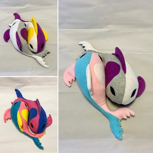 Op de afbeelding: Twee pluche vis speelgoedjes, een grijs met paarse en witte accenten, de andere blauw met roze en witte accenten, zijn in elkaar verstrengeld in de vorm van een hart.
