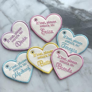 May include: Six heart-shaped fabric tags with embroidered edges and text. Each tag features the phrase If lost, please return to: followed by a name in cursive script. The tags have a small metal eyelet and come in pink, purple, blue, and yellow.