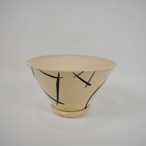 May include: A cream-colored ceramic planter with black abstract lines. The planter has a wide, shallow bowl shape and a smaller base.