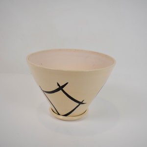 Puede incluir: Un macetero de cerámica de color crema con un diseño geométrico negro. El macetero tiene una abertura ancha y acampanada y una base estrecha.