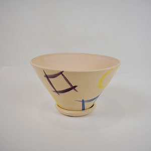 May include: A cream-colored ceramic planter with a wide, flared opening and a smaller base. The planter is decorated with abstract geometric shapes in yellow, blue, and purple.