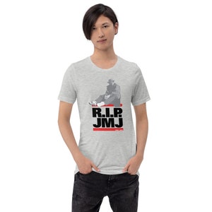 May include: A light gray t-shirt featuring a graphic of a person in a hat and coat, with the text "R.I.P. JMJ" in black and red. The shirt also includes the years "1965-2023".