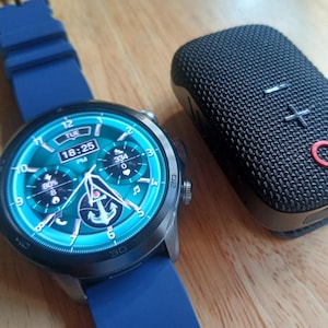 Op de afbeelding: Een zilveren smartwatch met een blauwe band toont de tijd 18:25 uur. De wijzerplaat is turquoise met witte en blauwe accenten. Een zwarte, rechthoekige luidspreker met een mesh textuur en rode en witte details ligt naast het horloge.