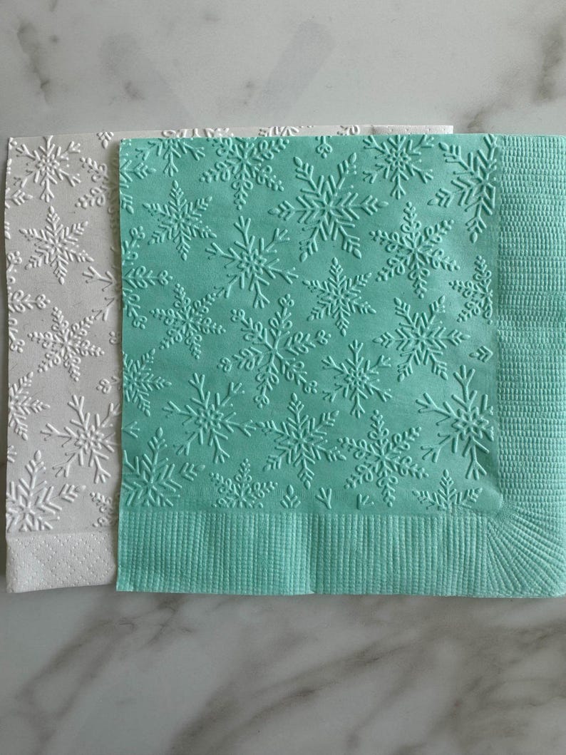 Puede incluir: Dos servilletas de papel con dise&ntilde;os de copos de nieve en relieve. Una servilleta es blanca y la otra de color verde azulado claro. La servilleta verde azulado tiene un borde texturizado. Adecuadas para eventos de invierno o festivos.